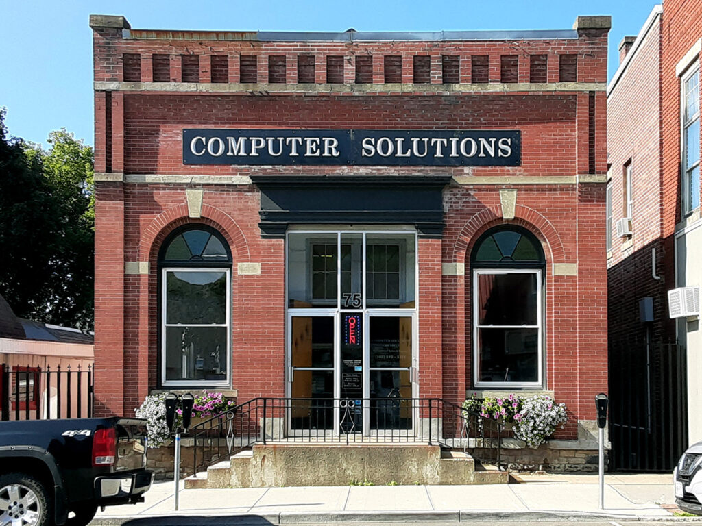 A red brick building labeled "Computer Solutions," featuring arched windows, a centered entrance with steps, and planters with flowers on either side. 